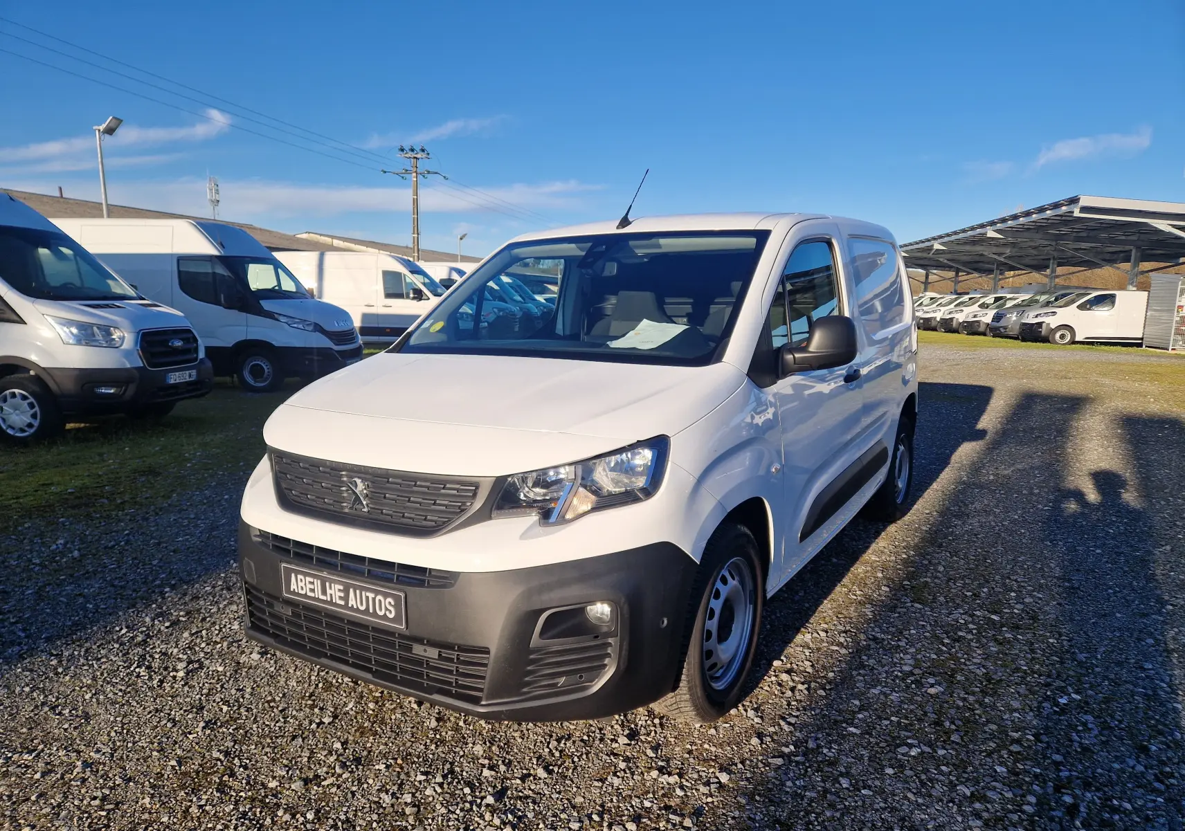 Vue 3/4 avant droite d’un Peugeot Partner blanc 2021 avec pare-chocs noir et calandre Peugeot, stationné sur un sol gravillonné.