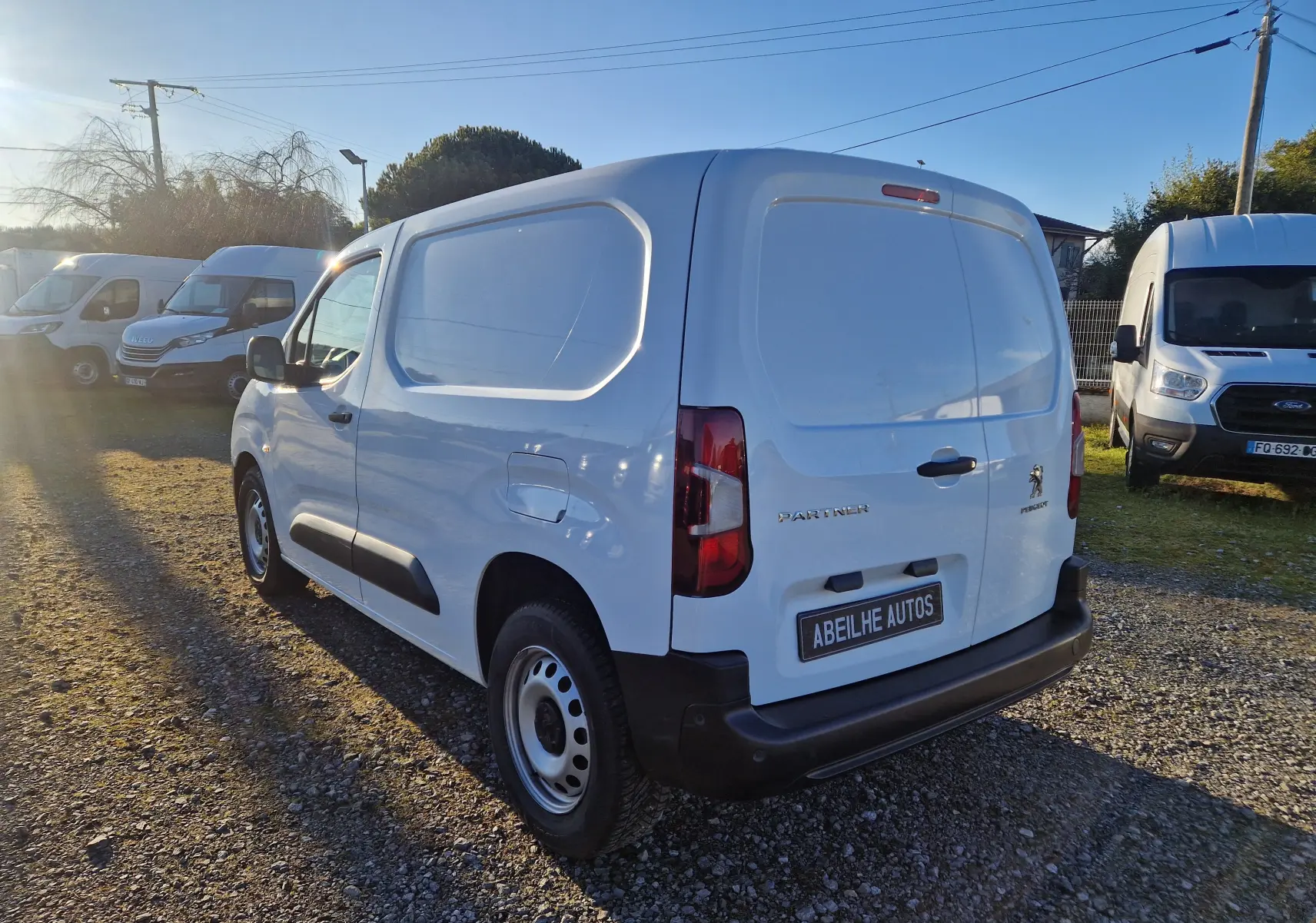 Vue 3/4 arrière droite d’un Peugeot Partner blanc 2021 utilitaire avec plaque Abeilhe Autos sur un terrain extérieur.
