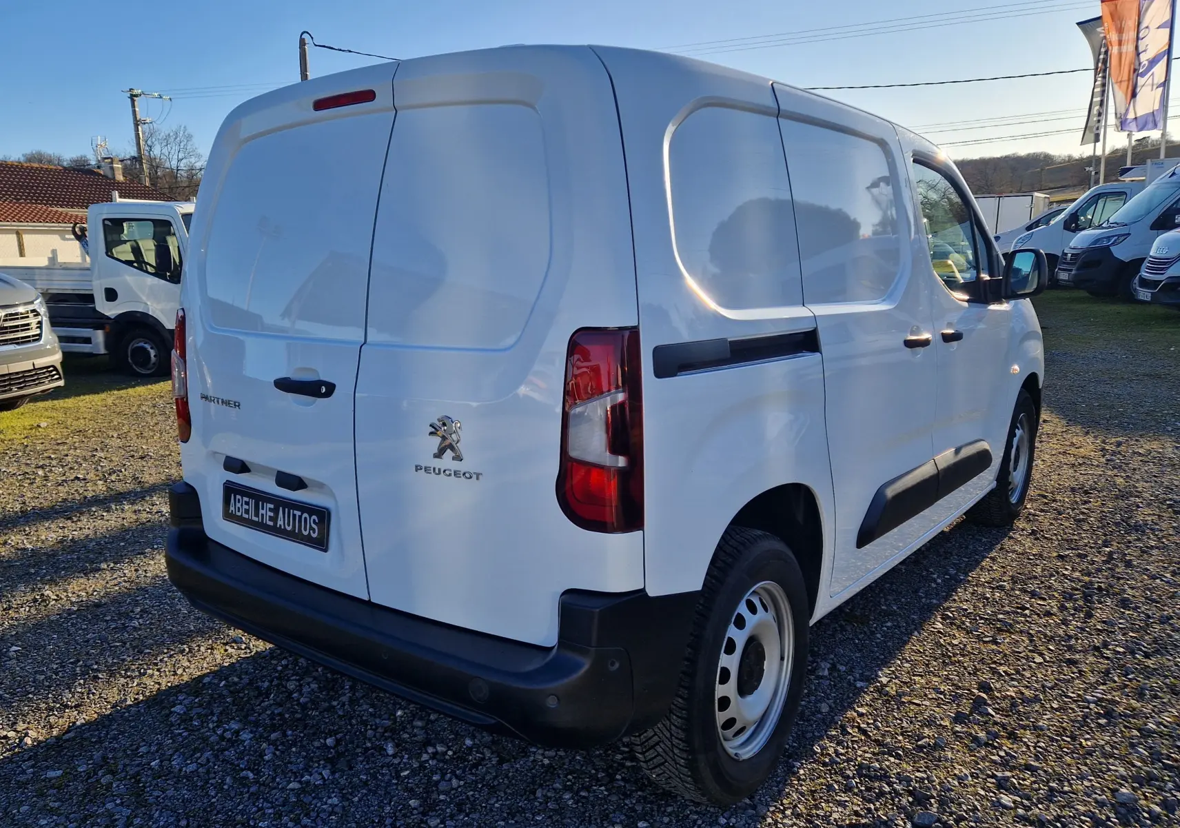 Vue 3/4 arrière droite d’un utilitaire Peugeot Partner blanc, version Standard 1.5 BlueHDi 100 Premium, sur terrain gravillonné.