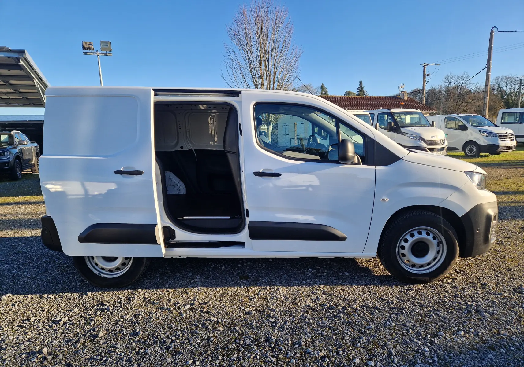 Vue de profil côté droit du Peugeot Partner blanc 2021 avec porte latérale coulissante ouverte sur un parking extérieur.