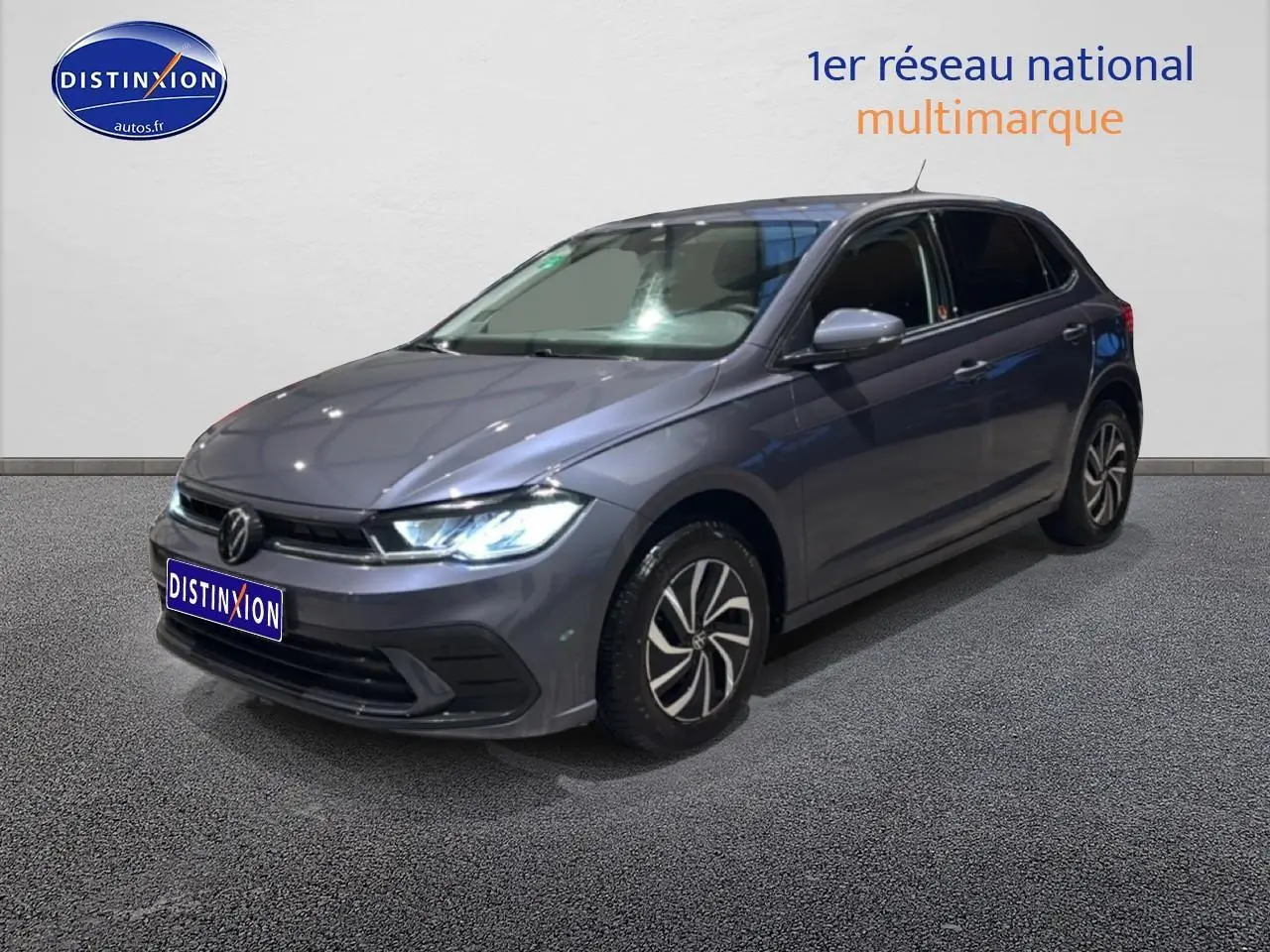 Volkswagen Polo gris cendre vue 3/4 avant droit avec phares allumés sur fond neutre intérieur.
