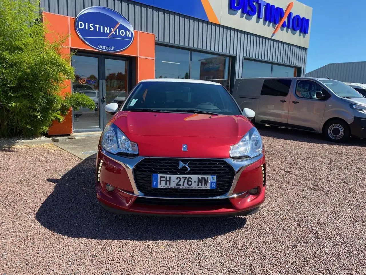 Vue avant d'une DS3 rouge à toit blanc, avec calandre chromée et phares distinctifs, stationnée devant un concessionnaire.