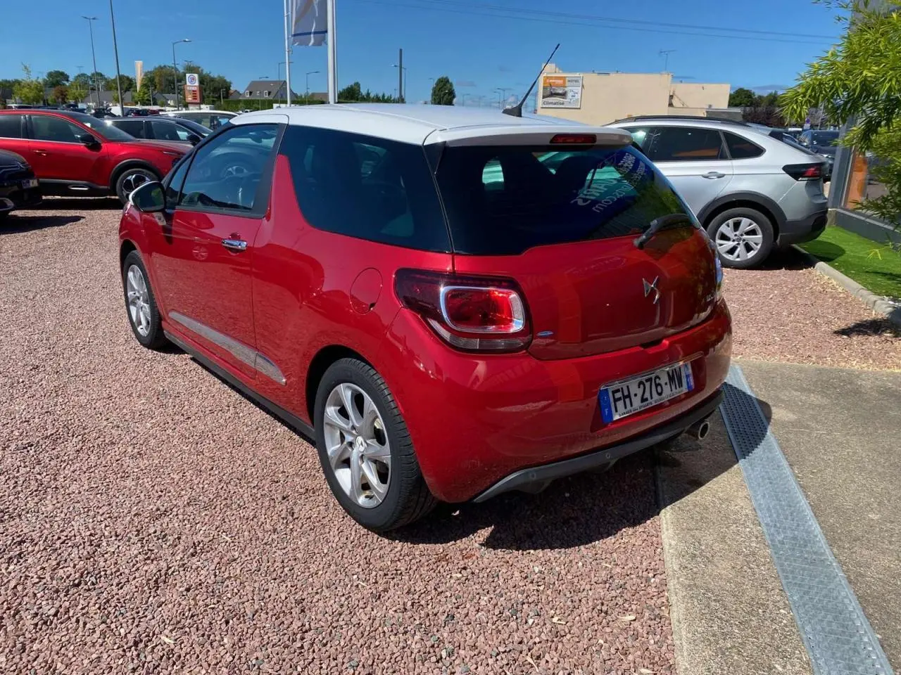 Vue 3/4 arrière droite d'une DS3 rouge à toit blanc, garée sur un sol en gravier sous un ciel dégagé.