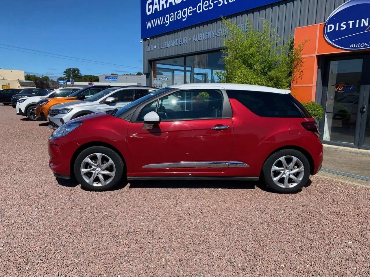 Vue de profil côté gauche d'une DS3 rouge à toit blanc, avec jantes alliage 16 pouces, stationnée devant un garage.