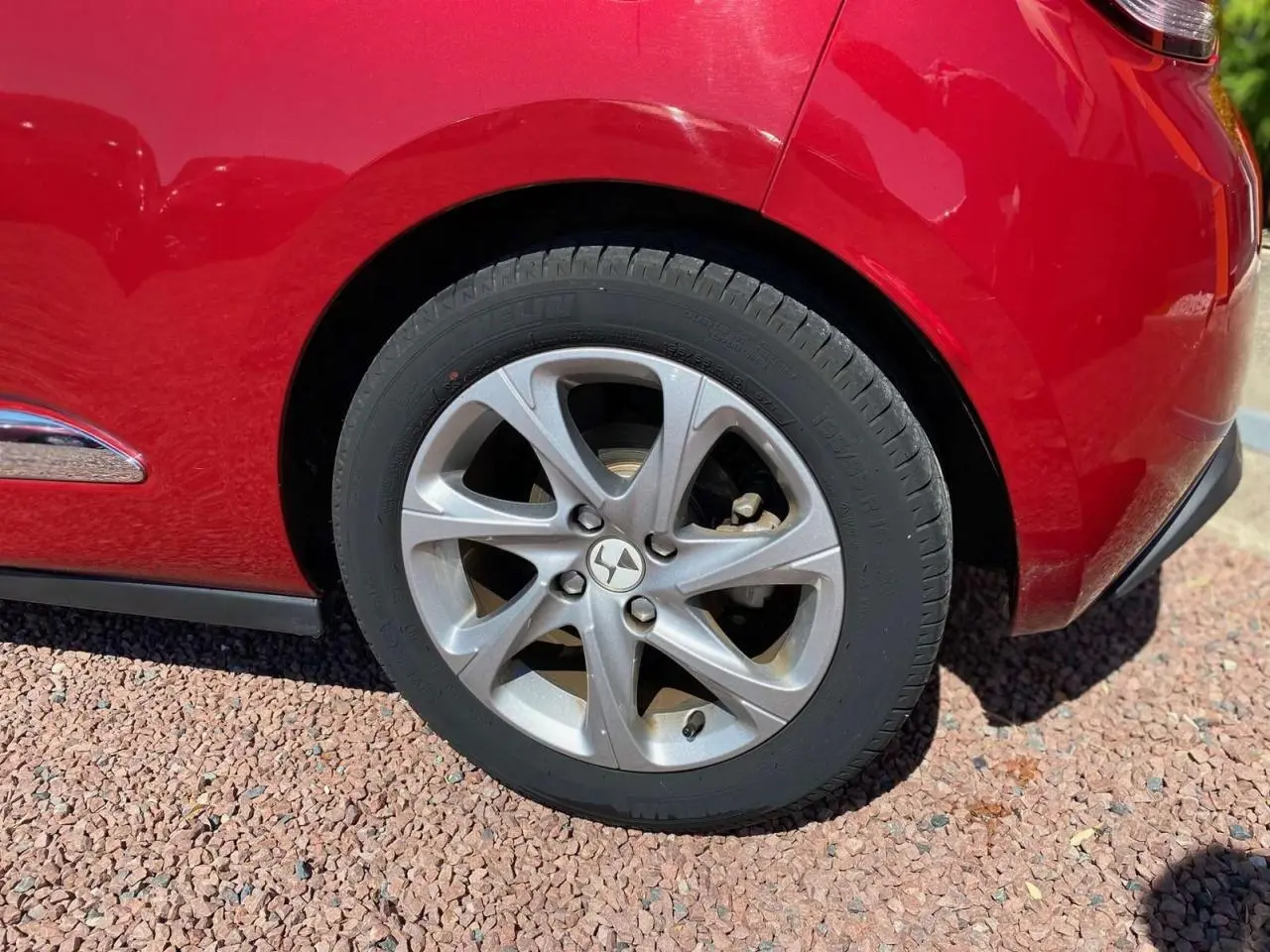 Gros plan sur la roue arrière gauche argentée d'une DS3 rouge avec toit blanc, sur sol gravillonné.