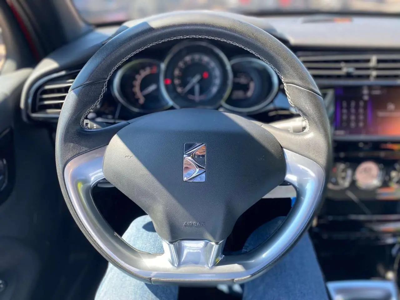 Vue rapprochée du volant DS3 en cuir noir avec logo DS, tableau de bord et écran tactile en arrière-plan flou.
