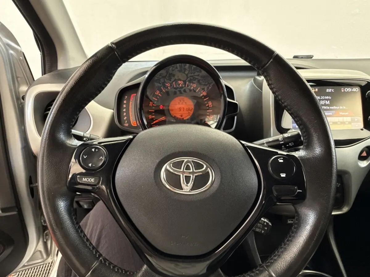 Vue intérieure centrée sur le volant noir de la Toyota Aygo gris clair, avec tableau de bord et écran multimédia visibles.