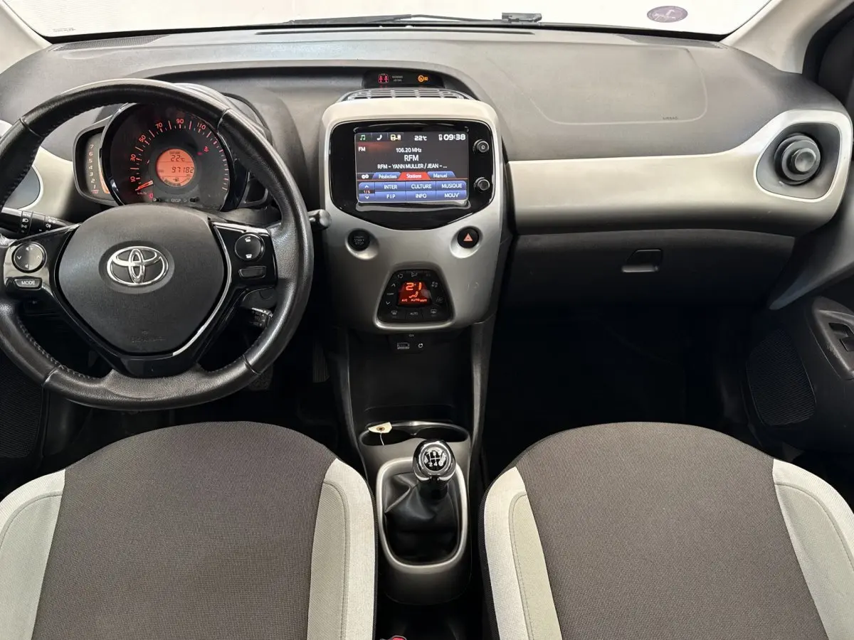Vue intérieure avant de la Toyota Aygo 2017, montrant le tableau de bord gris clair avec volant, écran tactile et levier de vitesses manuel.