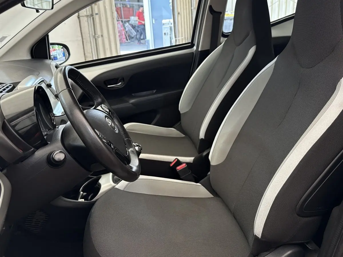 Vue intérieure côté conducteur de la Toyota Aygo gris clair, montrant les sièges avant et le volant noir.