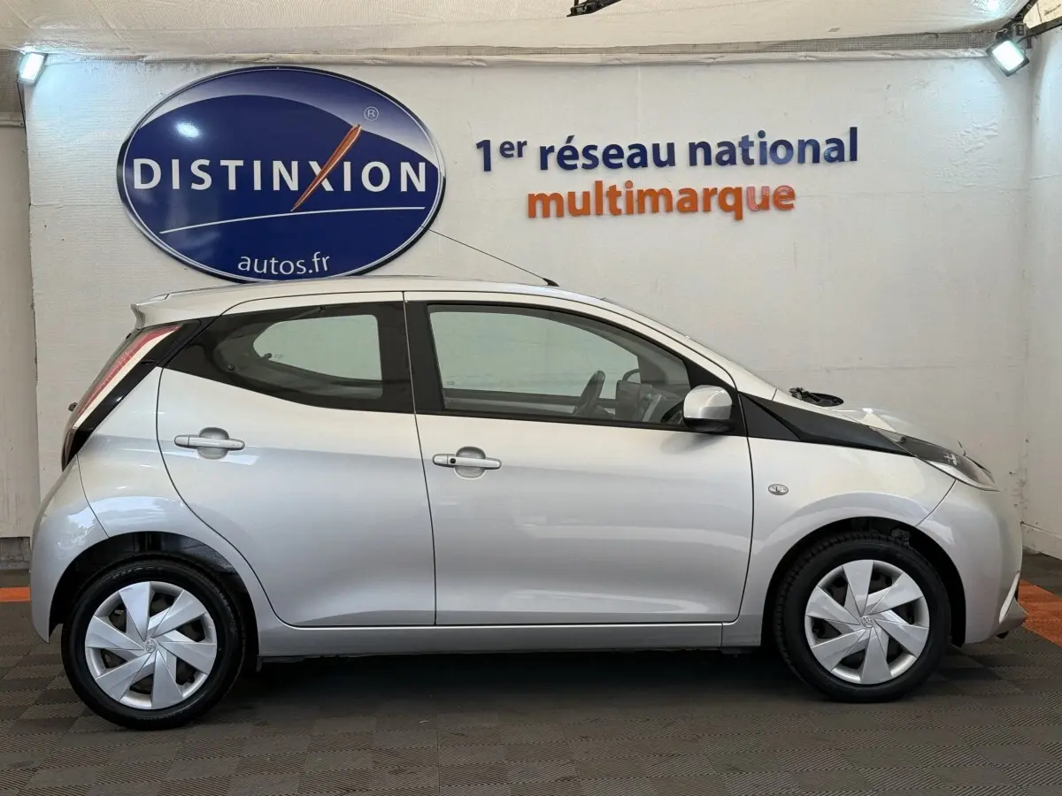 Profil latéral droit d'une Toyota Aygo gris clair 2017, avec jantes et rétroviseurs assortis, en intérieur showroom.