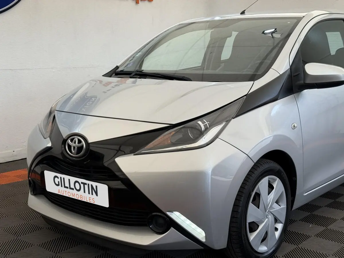 Toyota Aygo gris clair vue 3/4 avant droit, avec calandre noire distinctive et feux LED de jour allumés.