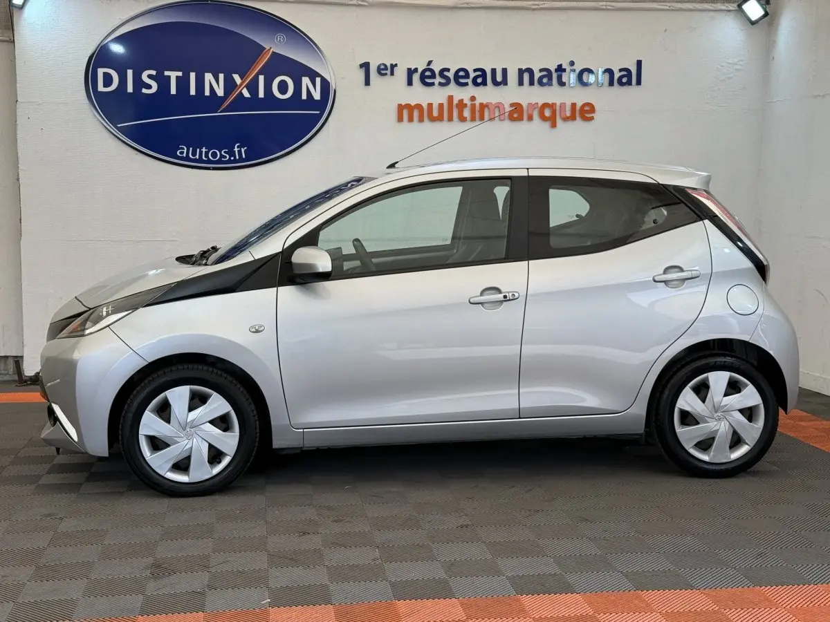 Profil droit d'une Toyota Aygo gris clair 2017 avec jantes argentées et bande noire distinctive sur l'aile avant.