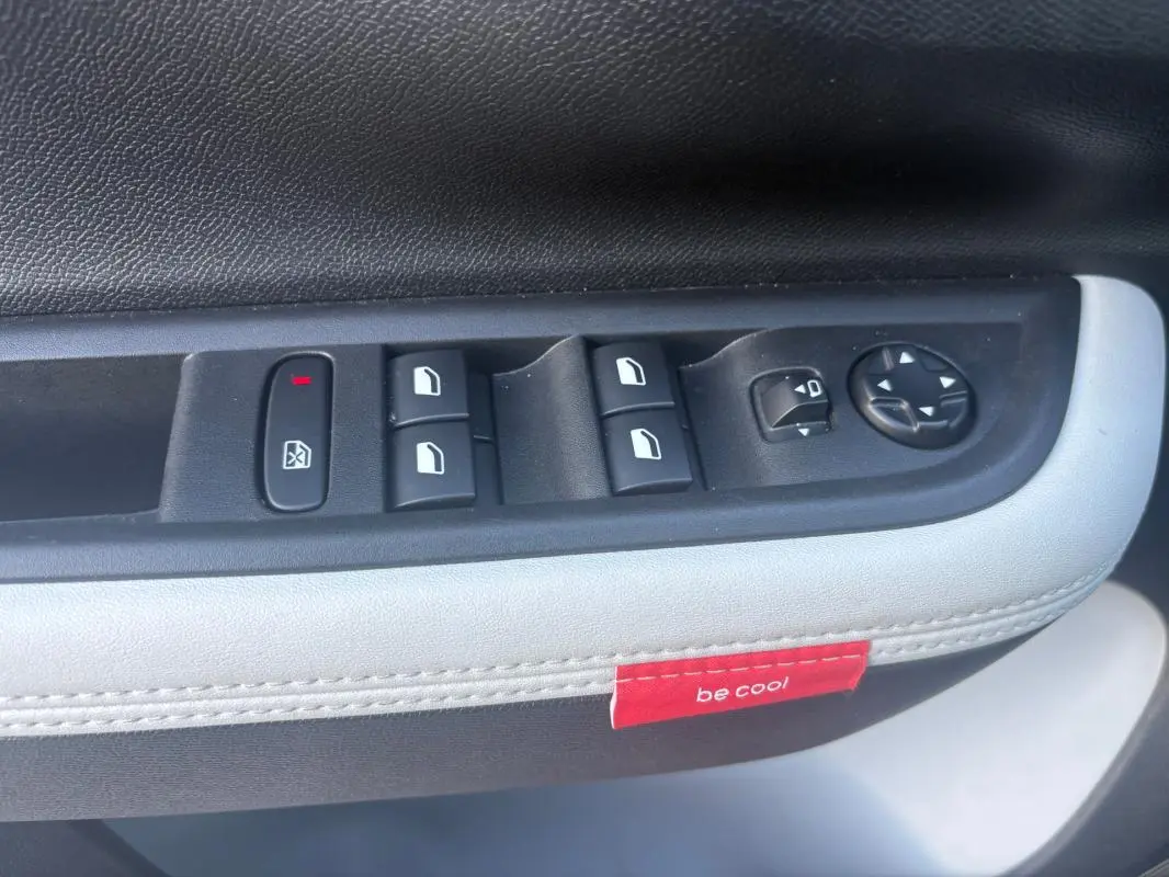 Gros plan sur la commande des vitres et rétroviseurs côté gauche avec surpiqûres blanches et étiquette rouge "be cool" dans une Citroën C3 gris foncé.