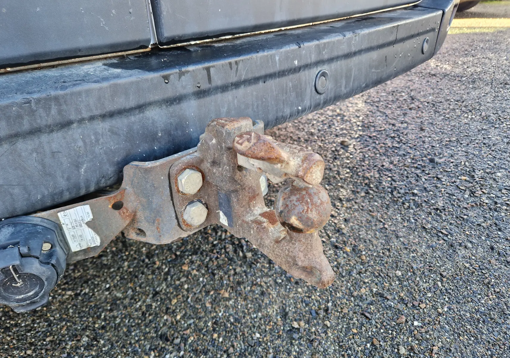 Gros plan sur l'attelage rouillé fixé au pare-chocs arrière noir d'un Renault Master blanc, vue latérale basse.