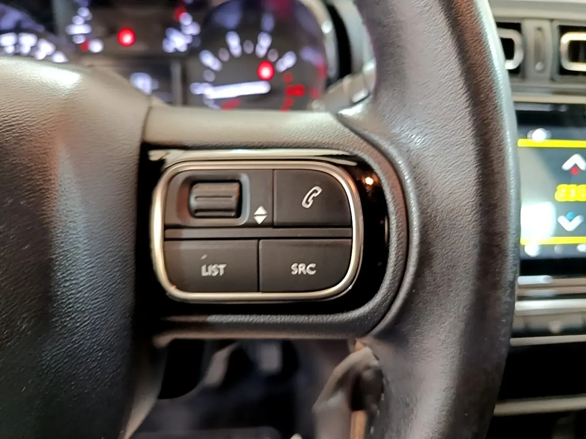 Gros plan sur les commandes gauche du volant cuir de la Citroën C3 blanche, avec tableau de bord et écran tactile en arrière-plan.