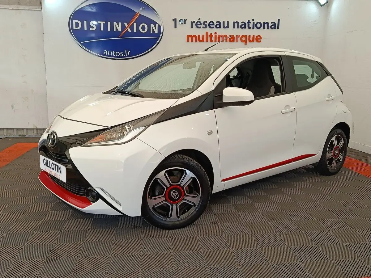 Vue 3/4 avant droite d'une Toyota Aygo blanche avec détails noirs et rouges, jantes stylisées et phares ellipsoïdaux.
