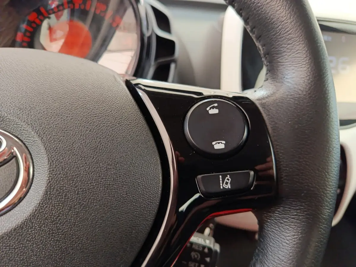 Gros plan sur le volant cuir noir de la Toyota Aygo blanc 2016, avec commandes téléphoniques et aide au maintien de voie.