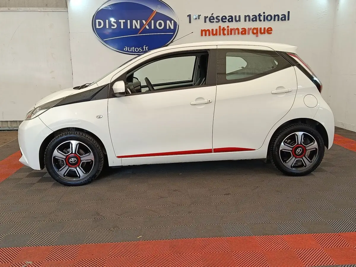 Profil côté gauche d'une Toyota Aygo blanche 2016 avec jantes noires et détails rouges, en intérieur showroom.
