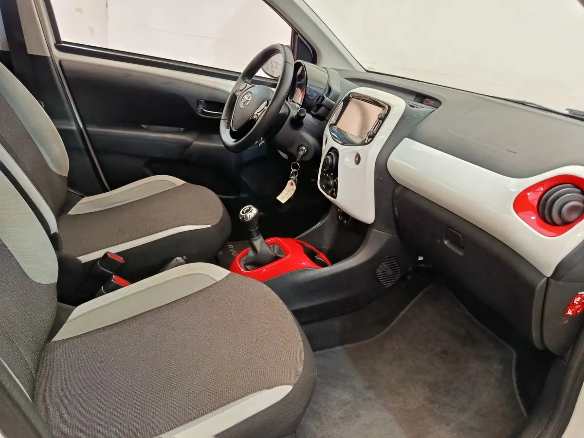 Vue intérieure côté conducteur de la Toyota Aygo blanche, avec tableau de bord bicolore et levier de vitesses manuel rouge.