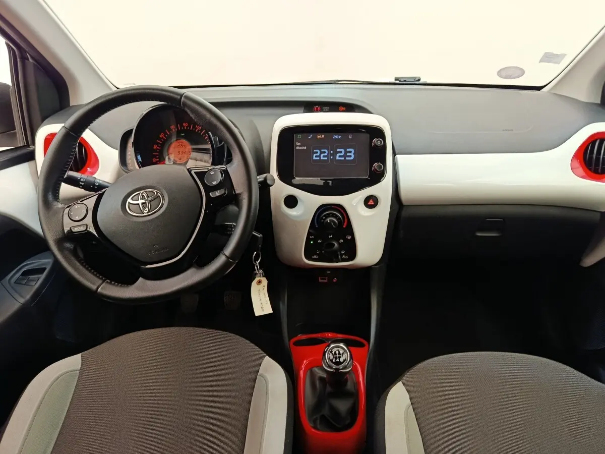 Vue intérieure frontale de la Toyota Aygo blanche, volant cuir, console centrale avec écran tactile et levier de vitesse rouge.