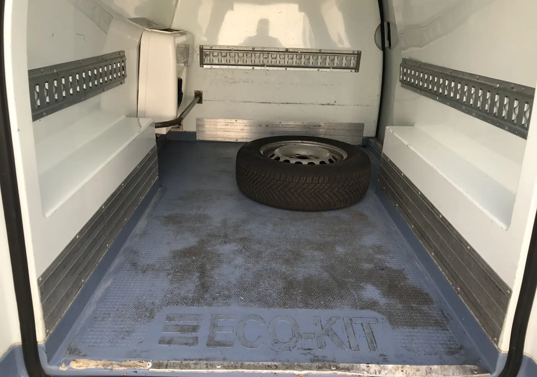 Vue intérieure arrière du Volkswagen Caddy Maxi Van frigo blanc de 2015 avec plancher bleu marqué ECO-KIT et roue de secours au centre.