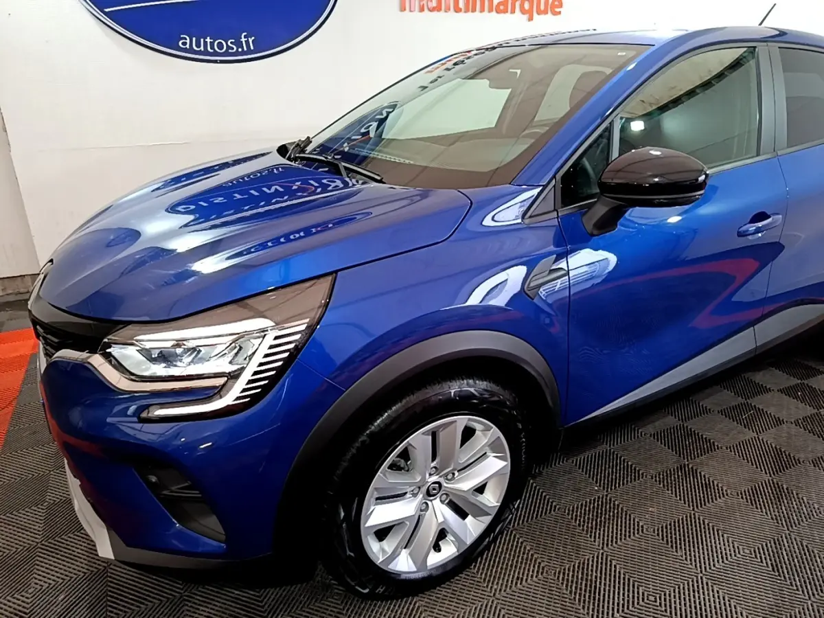 Renault Captur bleu en 3/4 avant droit, avec phares LED et jantes alliage visibles sur sol intérieur.