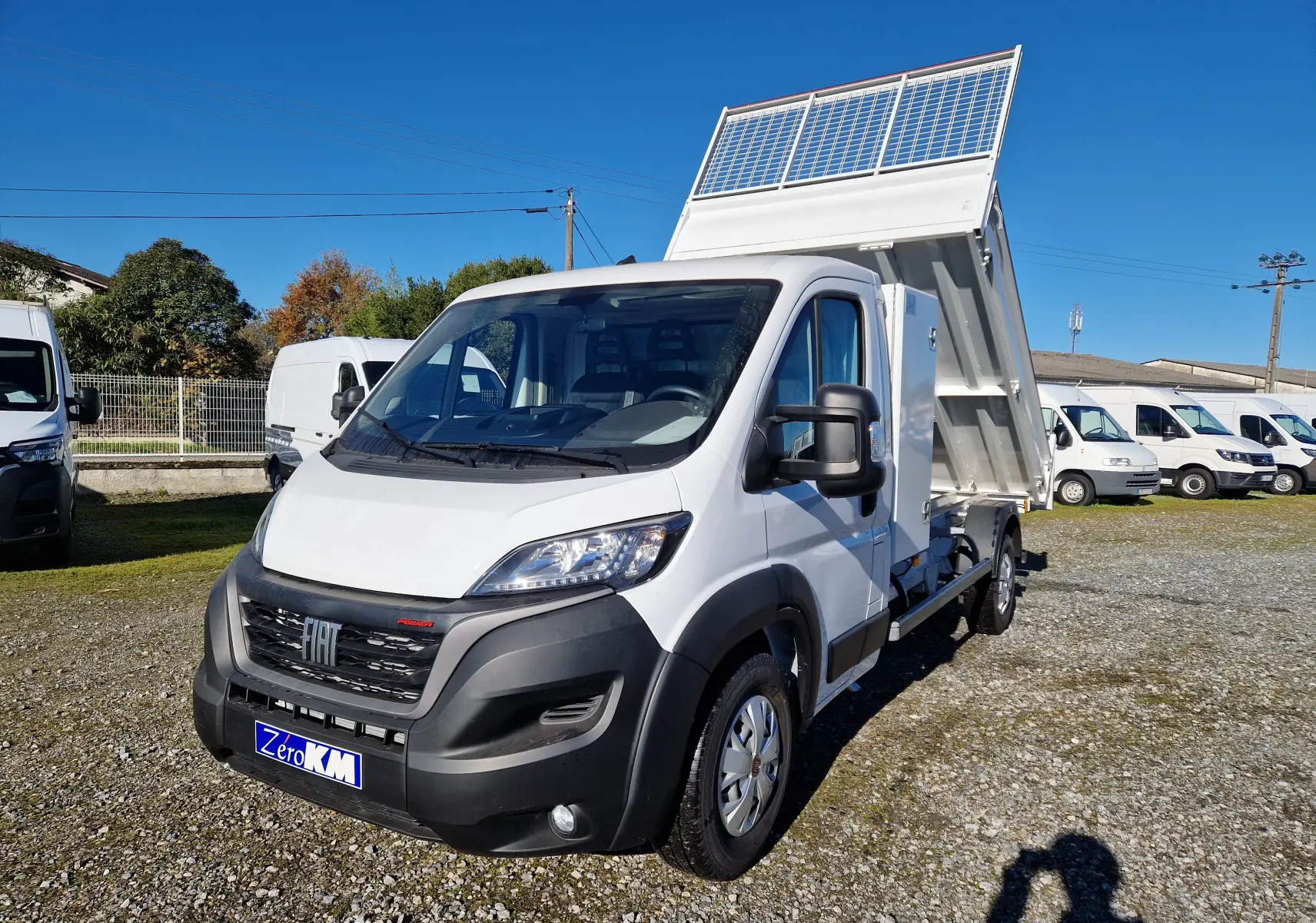 Fiat Ducato Chassis Benne blanc vu 3/4 avant droit avec benne acier relevée sur parking ensoleillé.