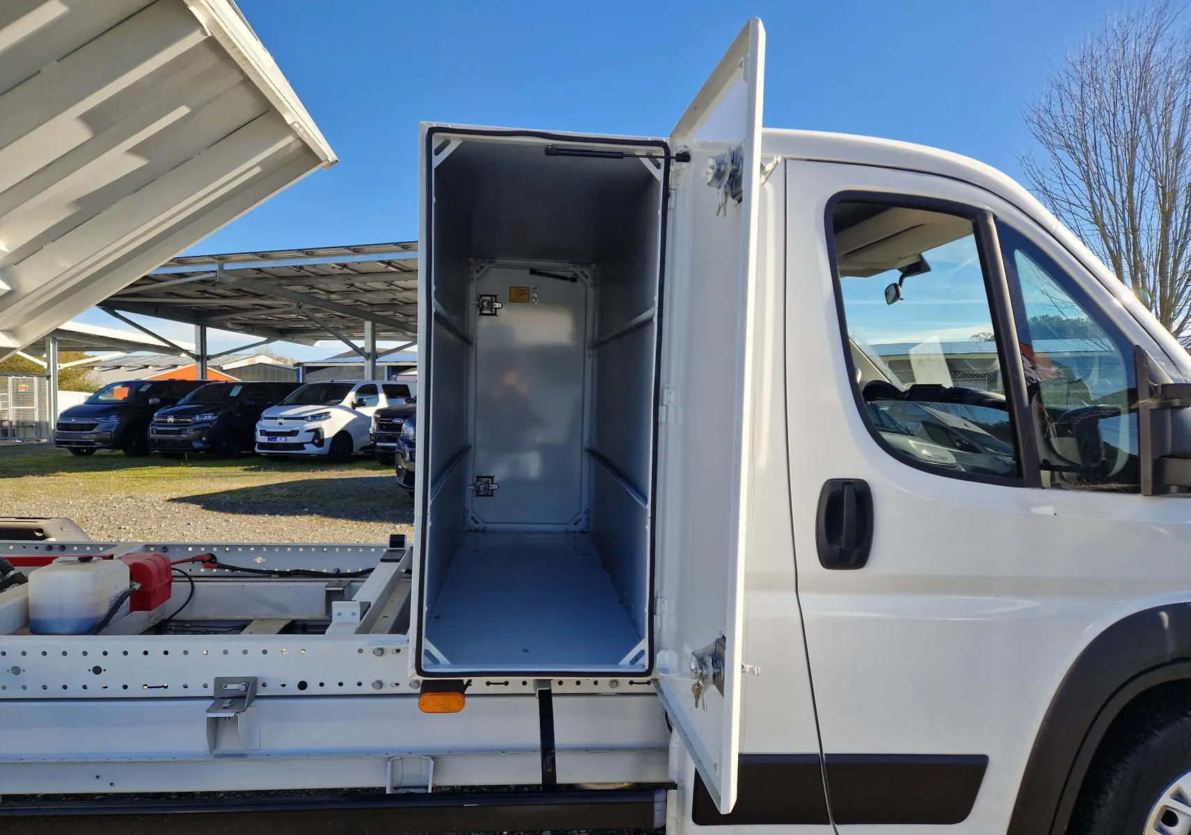 Vue latérale droite du FIAT Ducato Chassis Benne blanc 2023 avec coffre ouvert montrant l'intérieur métallique vide.