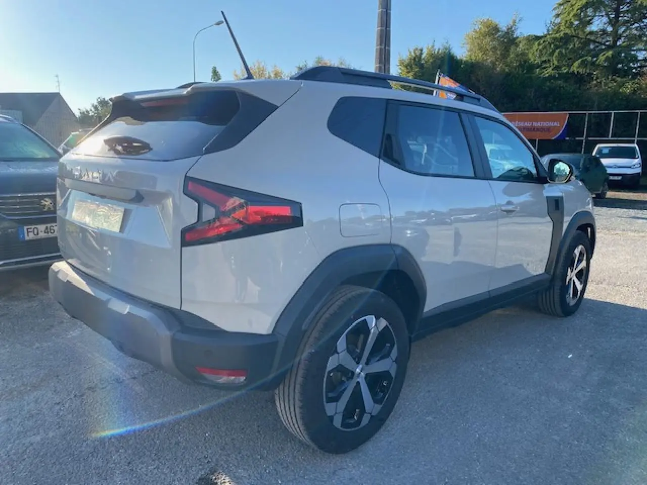 Vue 3/4 arrière droite du Dacia Duster 2025 beige Sandstone avec jantes alliage et barres de toit noires.