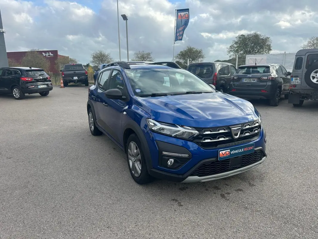 Vue 3/4 avant d'une Dacia Sandero Stepway bleu foncé avec barres de toit, garée sur un parking extérieur.