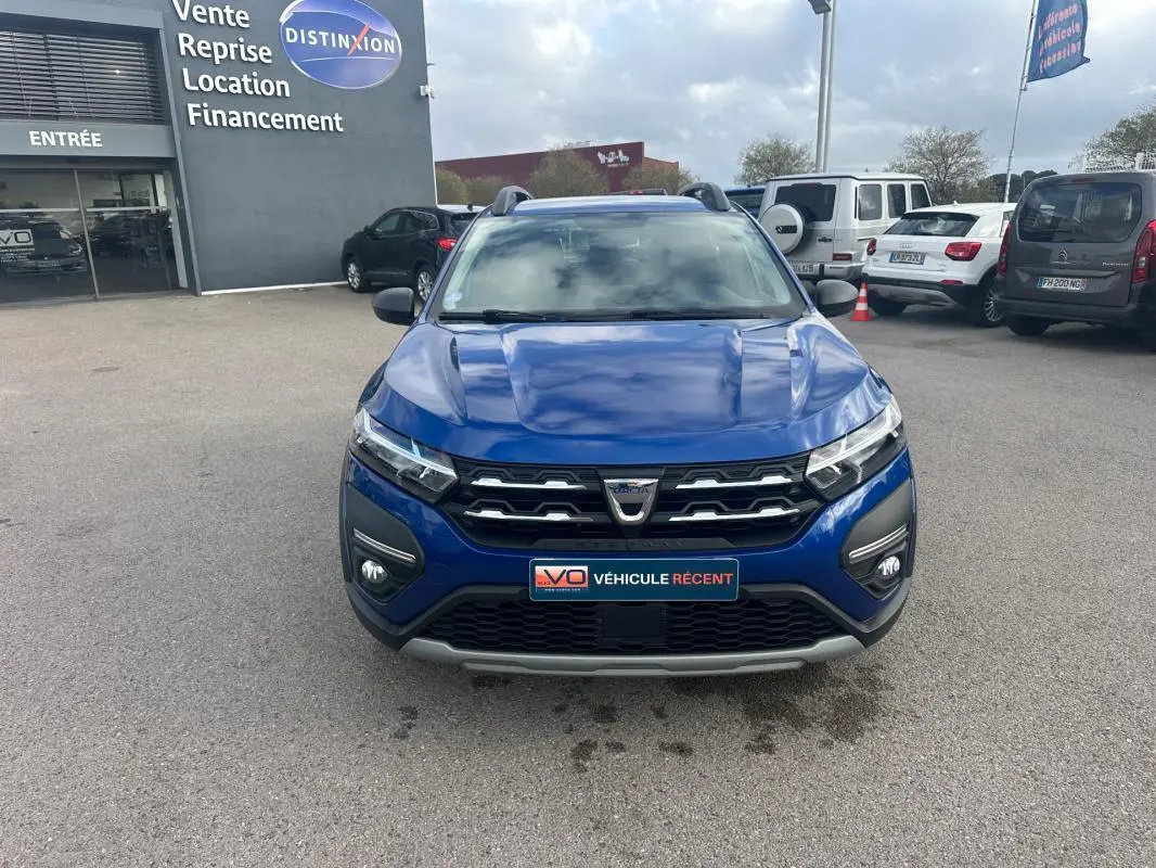 Vue frontale d'une Dacia Sandero Stepway bleu foncé avec barres de toit et feux allumés sur parking.