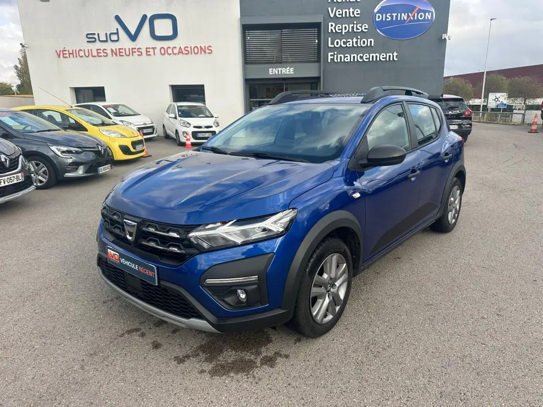 Vue 3/4 avant d'une Dacia Sandero Stepway bleu foncé avec barres de toit et phares allumés sur parking.