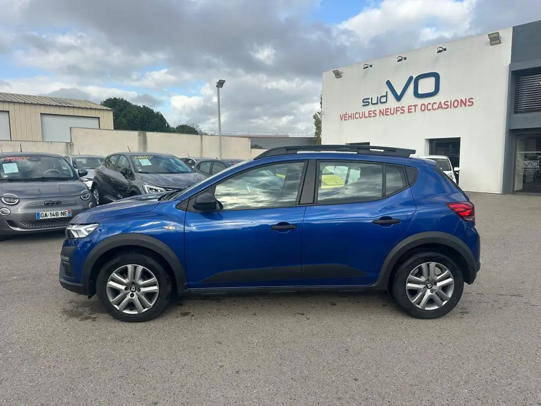Profil côté gauche d'une Dacia Sandero Stepway bleu foncé avec barres de toit et vitres surteintées, stationnée en extérieur.