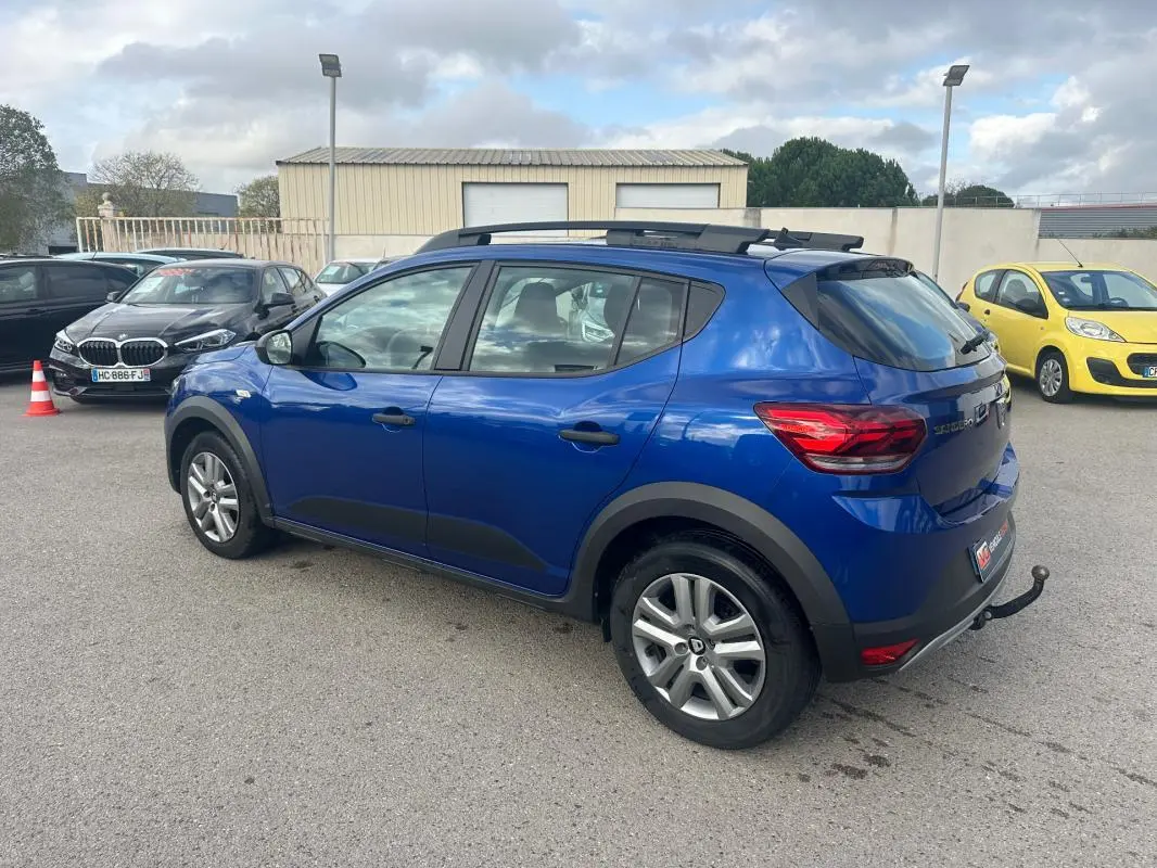 Vue 3/4 arrière gauche d'une Dacia Sandero Stepway bleu foncé avec barres de toit et attelage visible.