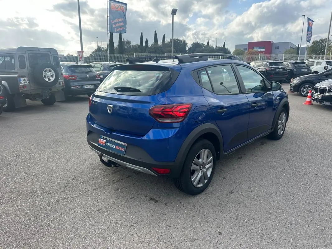 Vue 3/4 arrière droite d'une Dacia Sandero Stepway bleu foncé avec barres de toit et vitres surteintées dans un parking.
