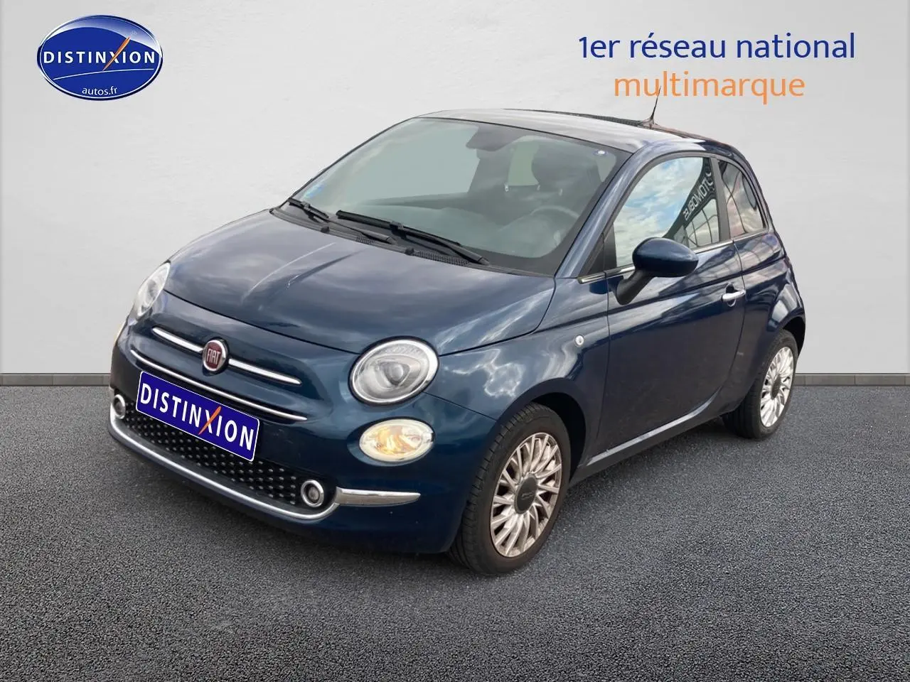 Vue 3/4 avant droite d'une Fiat 500 bleu foncé avec jantes alu et phares allumés sur fond neutre.