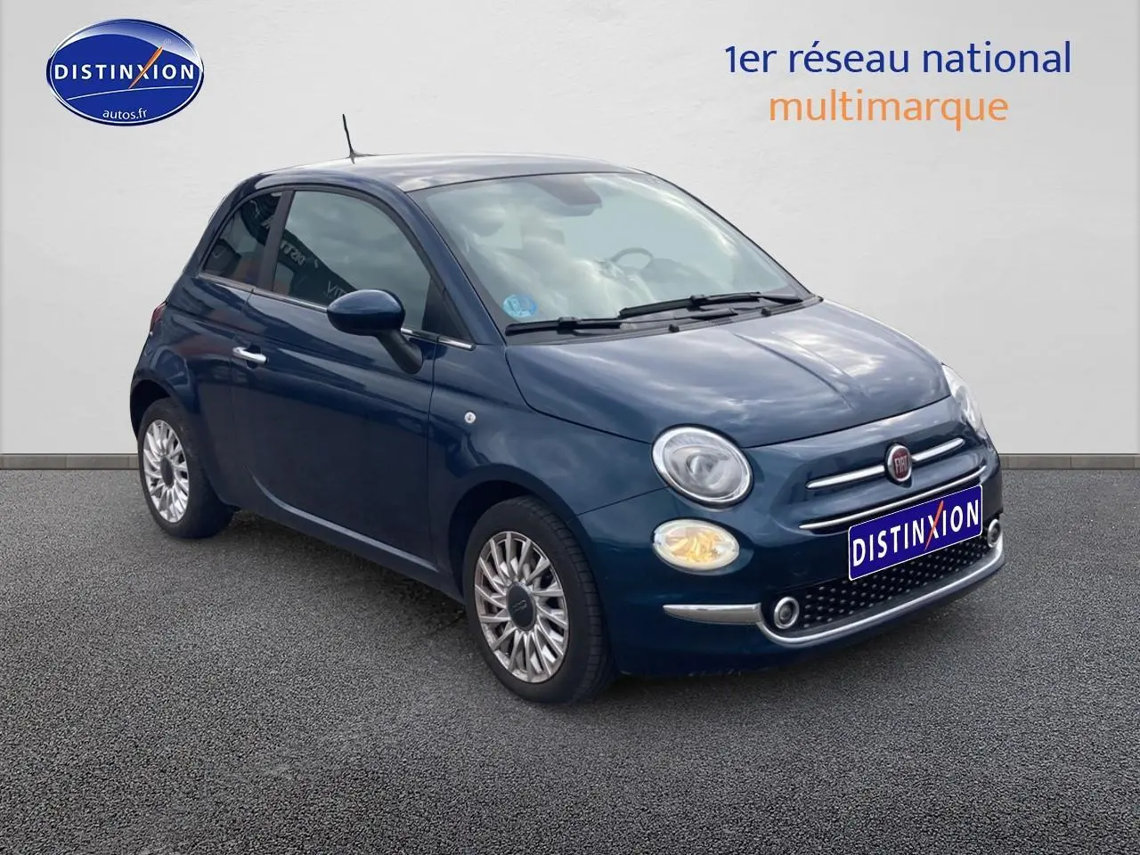 Vue 3/4 avant gauche d'une Fiat 500 bleu foncé avec jantes alu et phares allumés sur fond neutre.