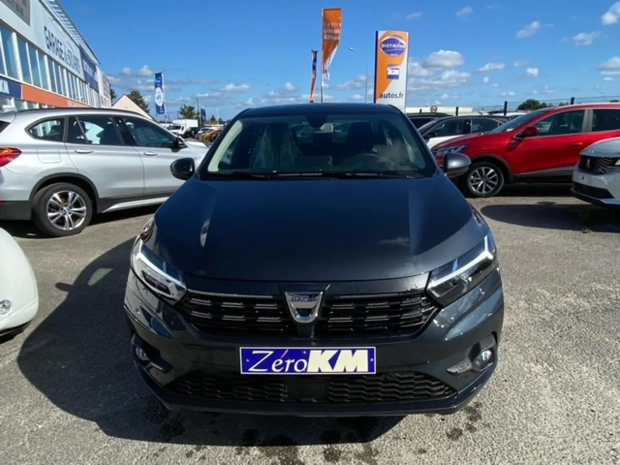 Vue frontale d'une Dacia Logan gris foncé 2022 avec calandre noire et plaque "Zero KM" sur un parking extérieur.