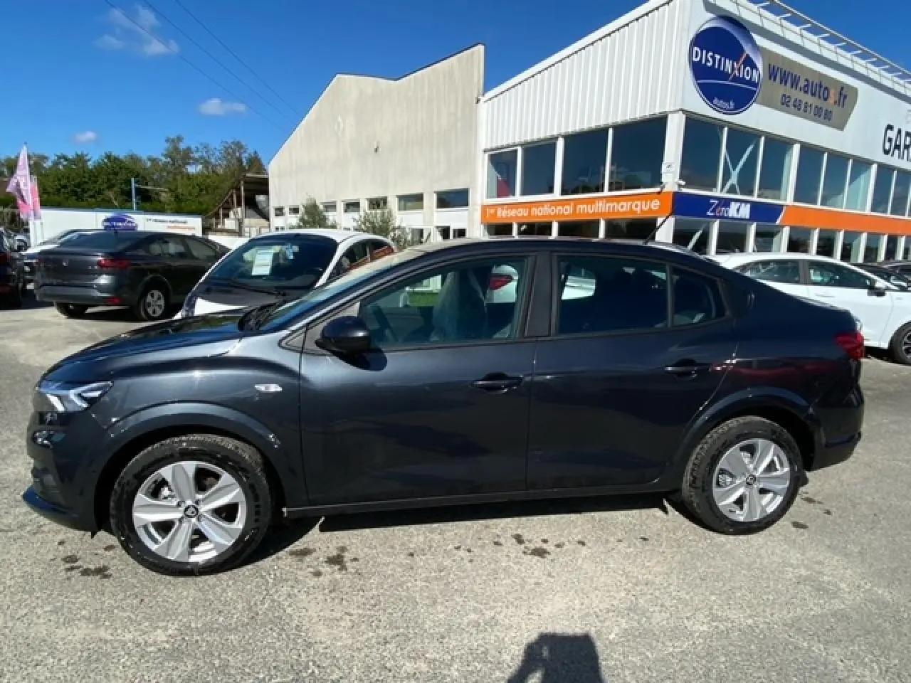 Vue de profil côté gauche d'une Dacia Logan gris foncé 2022 stationnée devant un garage multimarque.