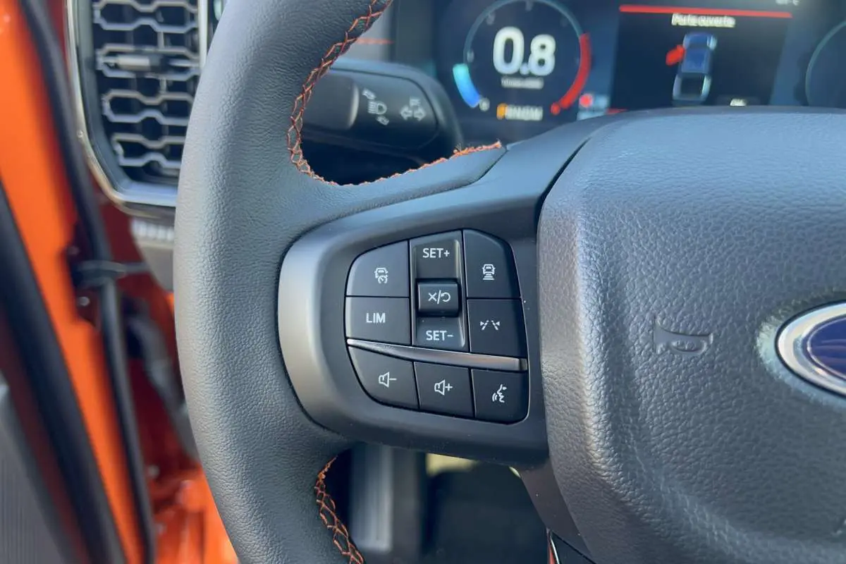 Gros plan sur le volant multifonctions noir avec surpiqûres orange du Ford Ranger Wildtrak Ignite orange, tableau de bord digital en arrière-plan.