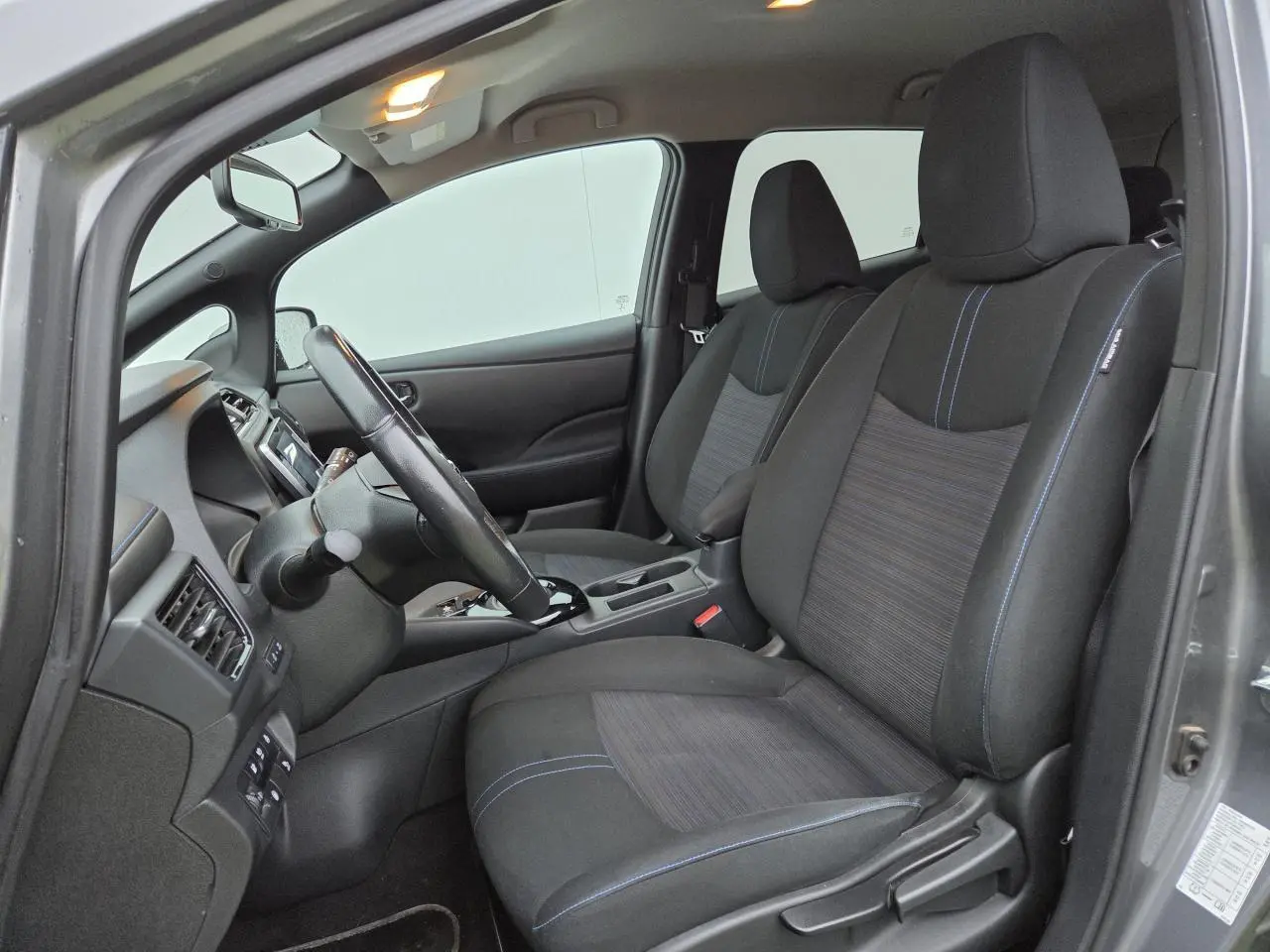 Vue intérieure côté conducteur de la Nissan Leaf II gris foncé, montrant les sièges noirs avec surpiqûres bleues et le volant cuir.