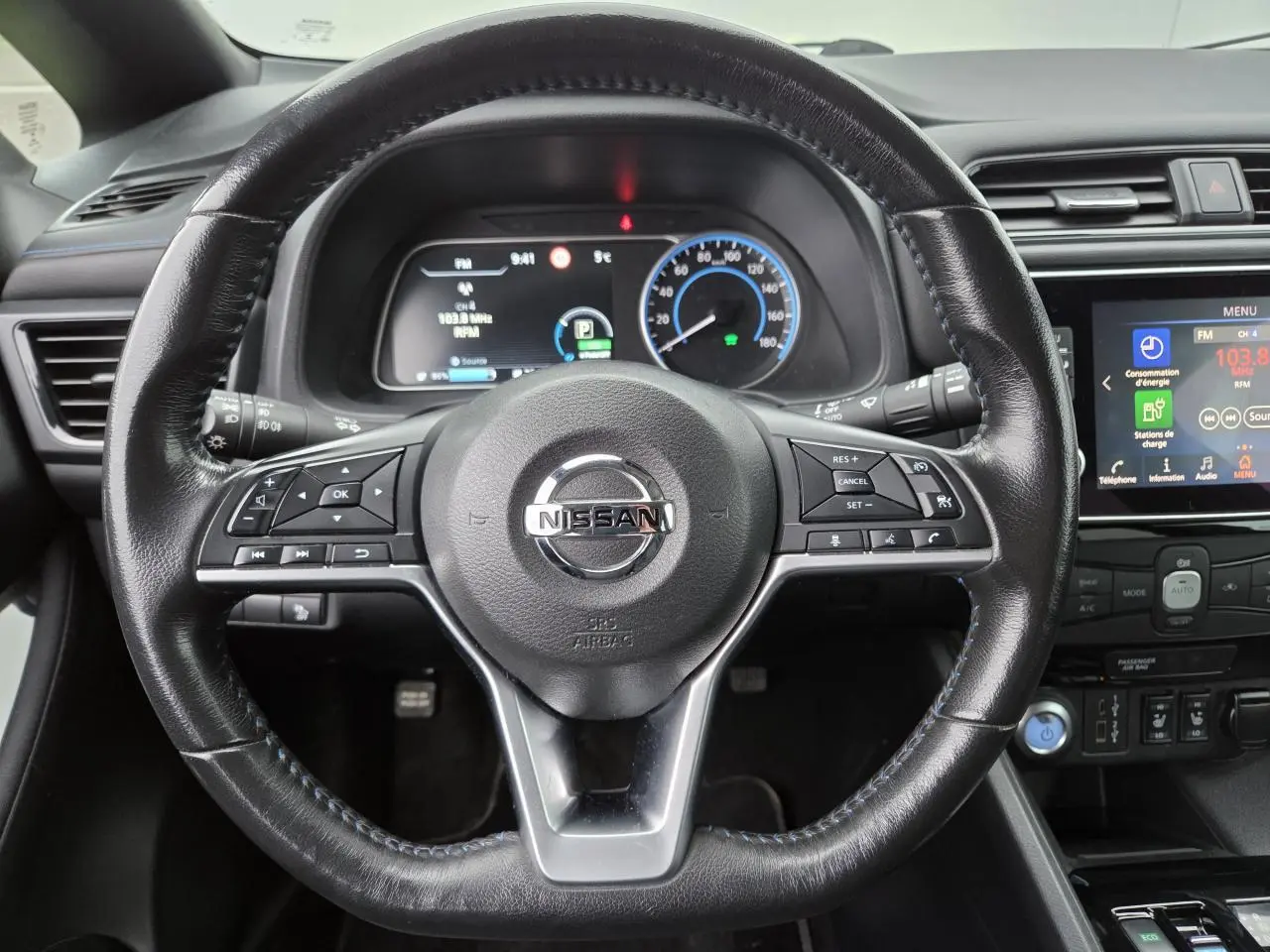 Vue intérieure centrée sur le volant cuir noir de la Nissan Leaf II 150CH avec tableau de bord digital et écran tactile.