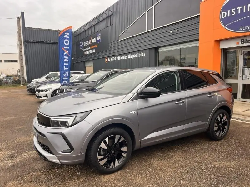 Vue 3/4 avant droite d'un Opel Grandland 2023 gris Kontrast métallisé avec jantes bi-ton et rétroviseurs noirs devant un bâtiment commercial.