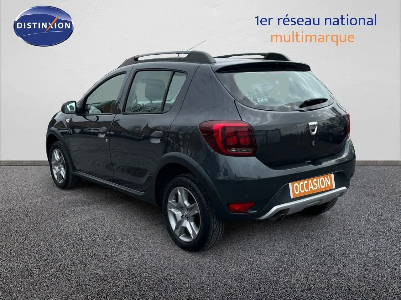 Vue 3/4 arrière droite d'une Dacia Sandero Stepway 0.9 TCe gris foncé de 2016 avec barres de toit et jantes alliage.