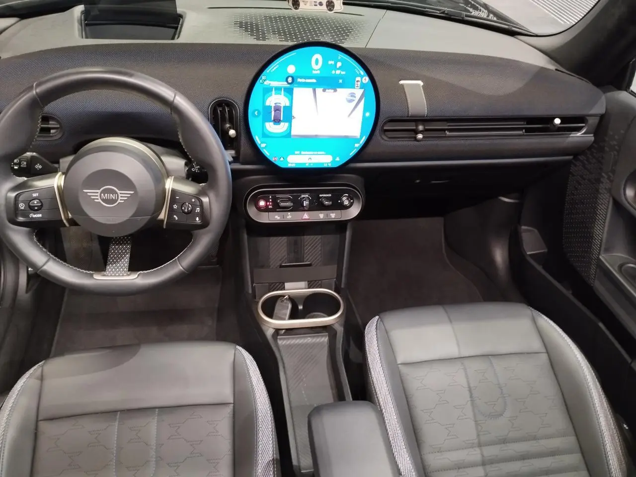 Intérieur avant noir du MINI Cabriolet 2025, vue centrée sur le volant et l'écran tactile circulaire lumineux.