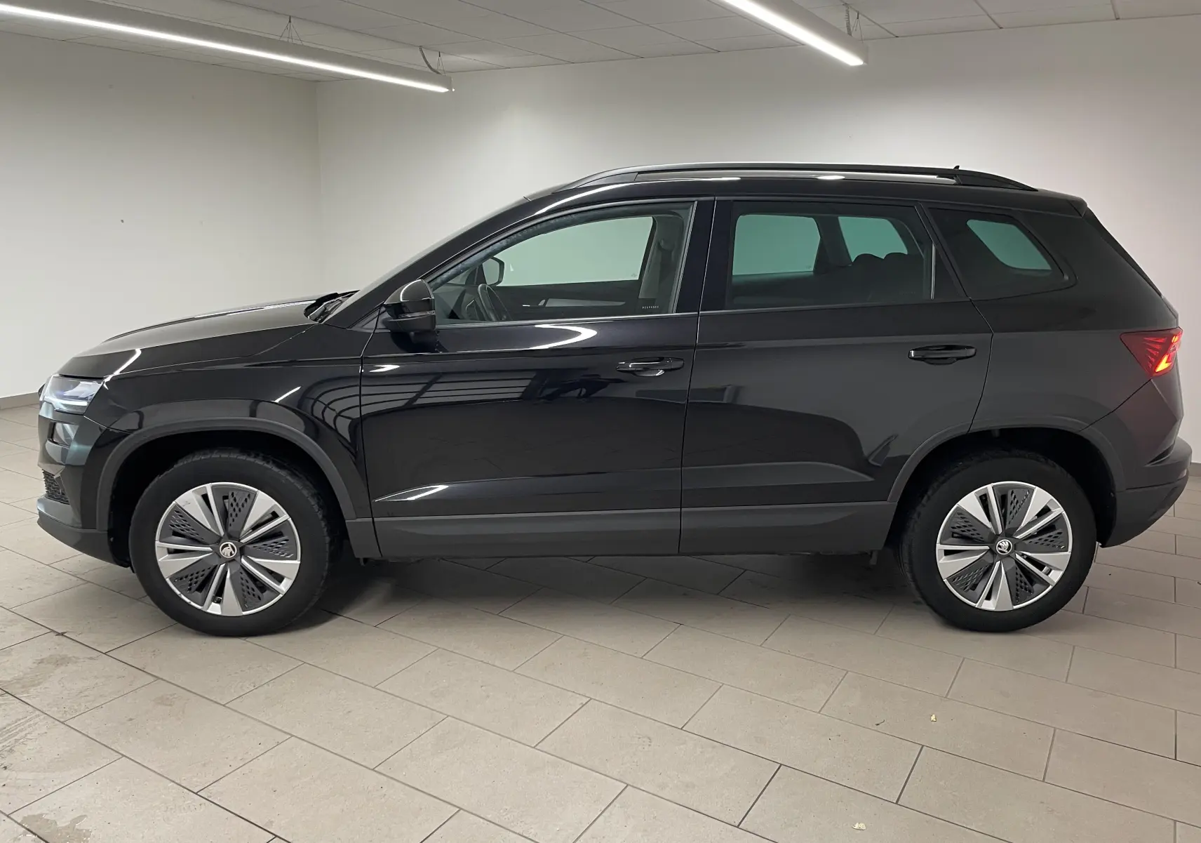 Profil côté gauche du SUV SKODA KAROQ 2023 en noir Magic nacré, avec jantes modernes et feux arrière allumés.