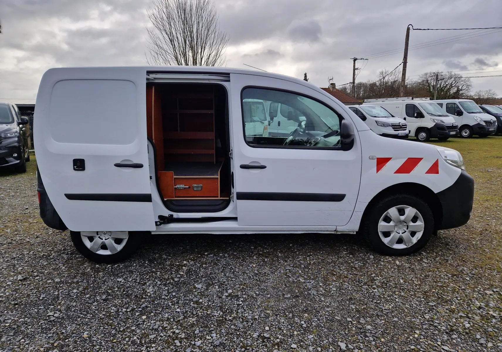 Vue de profil côté droit d’un Renault Kangoo blanc 2019 avec porte coulissante ouverte révélant des étagères en bois à l’intérieur.