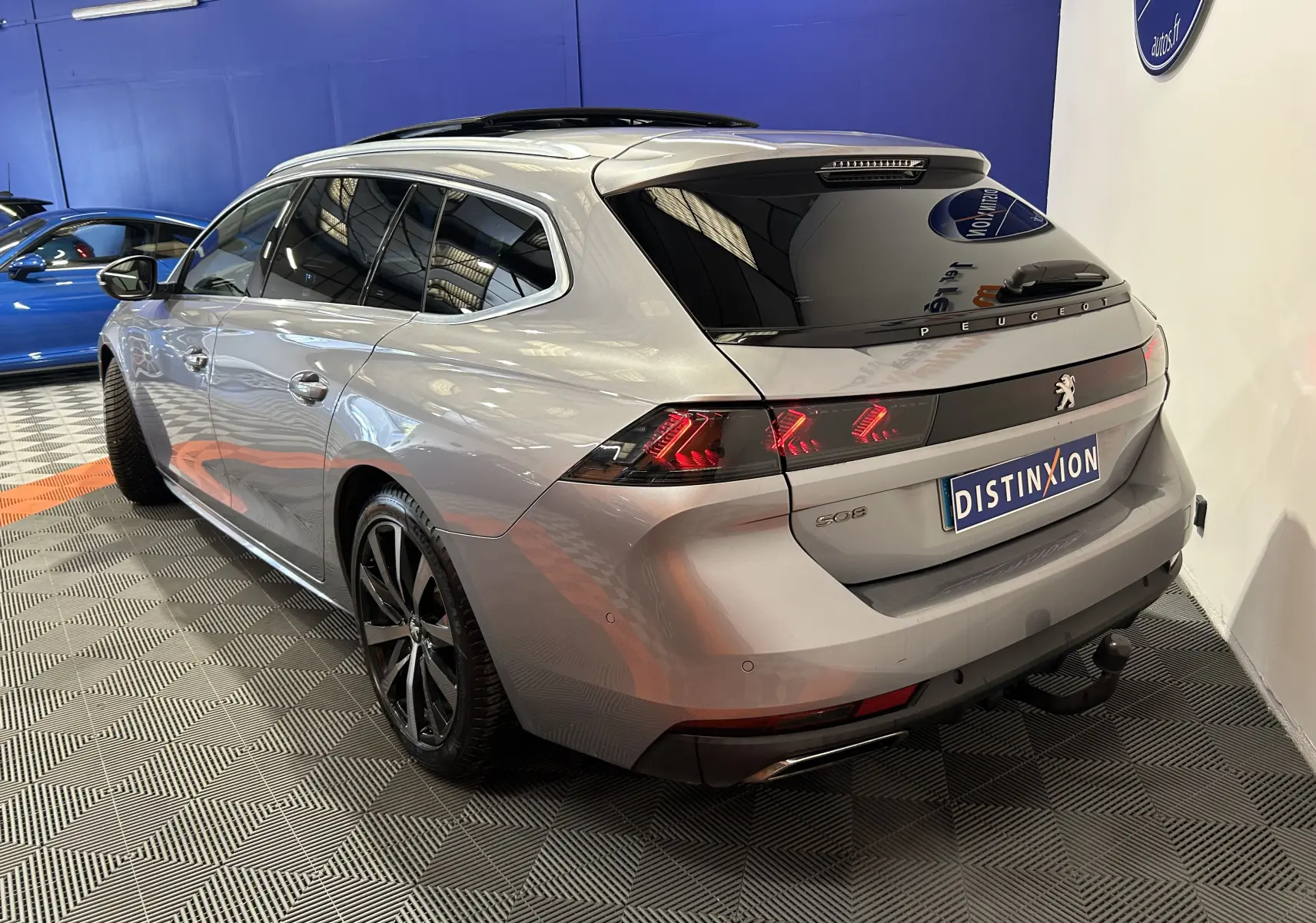 Vue 3/4 arrière droite d'une Peugeot 508 SW gris Platinium avec attelage et toit ouvrant dans un showroom.