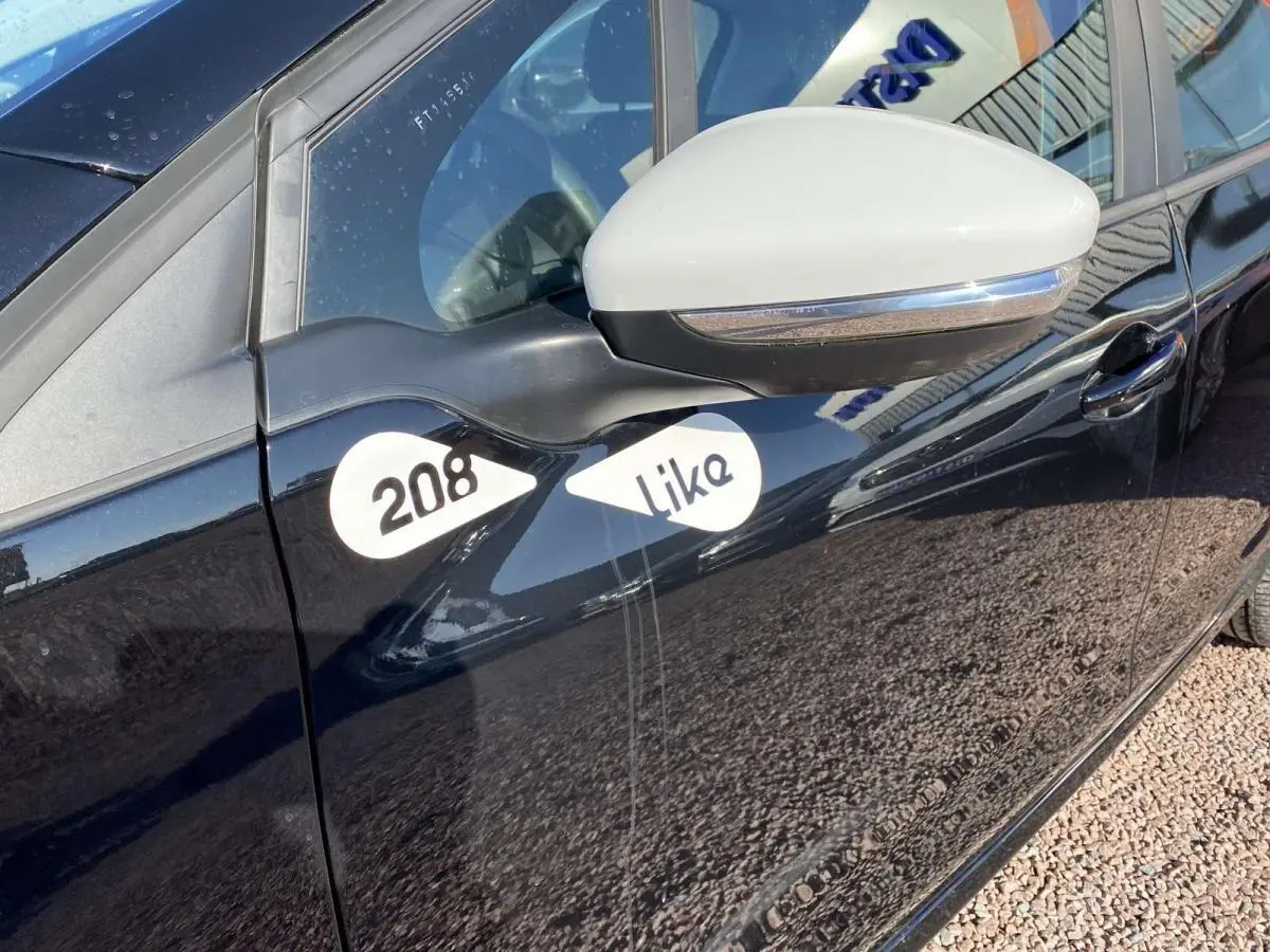 Vue latérale gauche rapprochée d’une Peugeot 208 noire avec rétroviseur blanc et logos 208 Like sur la porte avant.