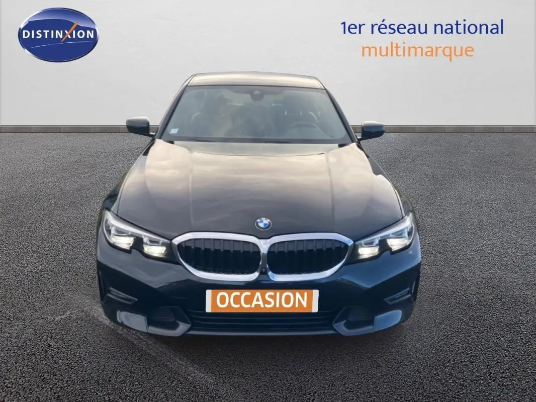 Vue avant d'une BMW Série 3 G20 noire de 2019 avec calandre chromée et phares allumés sur parking.